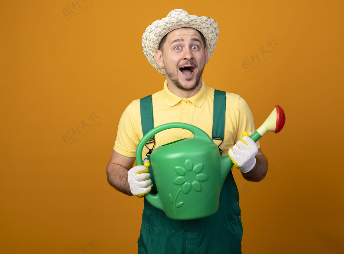 浇水年轻的园丁穿着连体衣 戴着帽子 手里拿着浇水罐 站在橙色的墙上 面带微笑地看着前面看人人