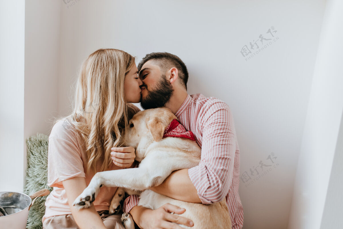 夫妇一对浪漫的情侣在阳光明媚的房间里亲吻穿着粉色衬衫的男人和他的女朋友拥抱着狗在一起家庭狗