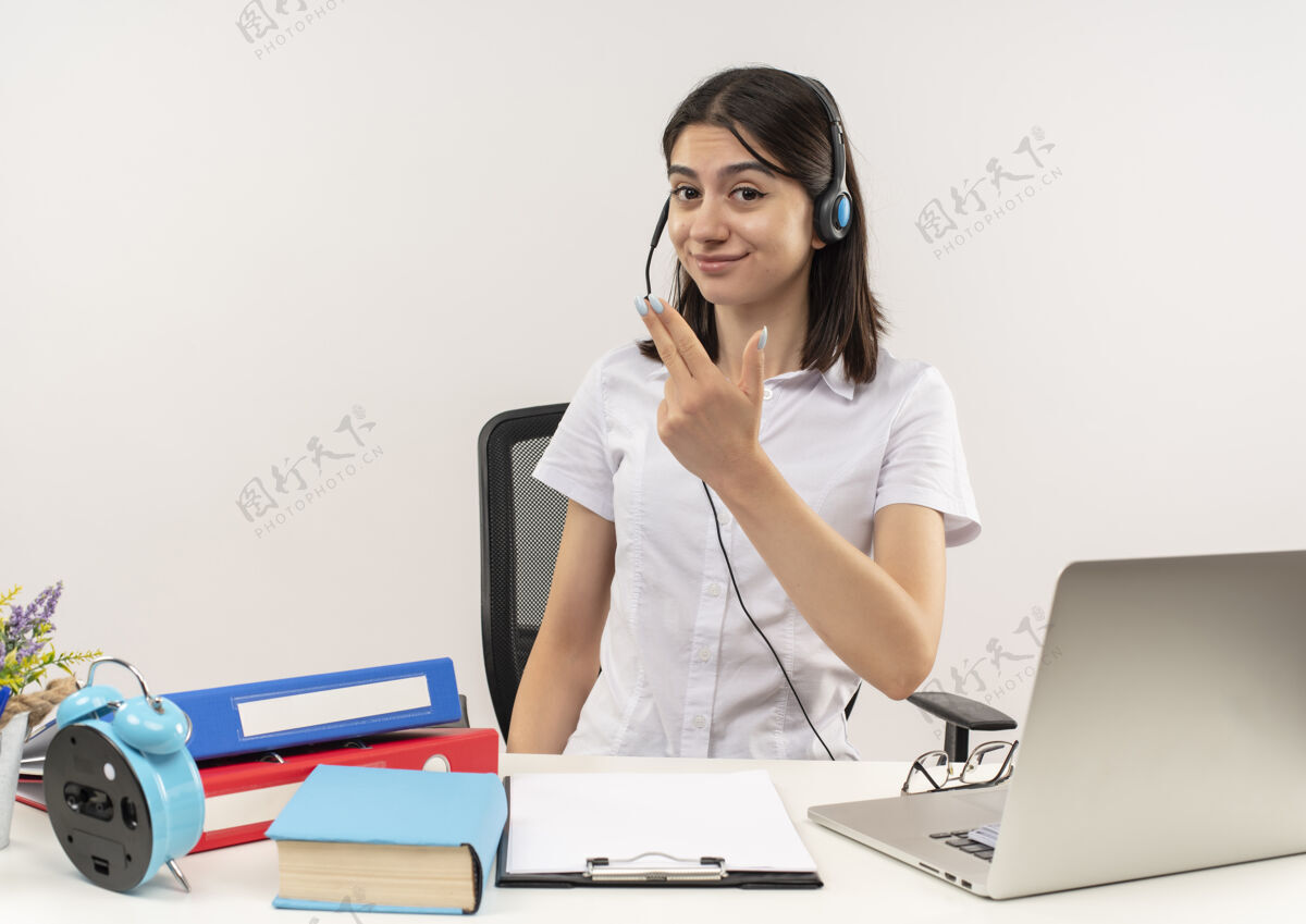 看穿着白衬衫戴着耳机的年轻女孩 两个手指面带微笑地看着前面 坐在桌子旁 白色的墙上放着文件夹和笔记本电脑桌子麦克风手指