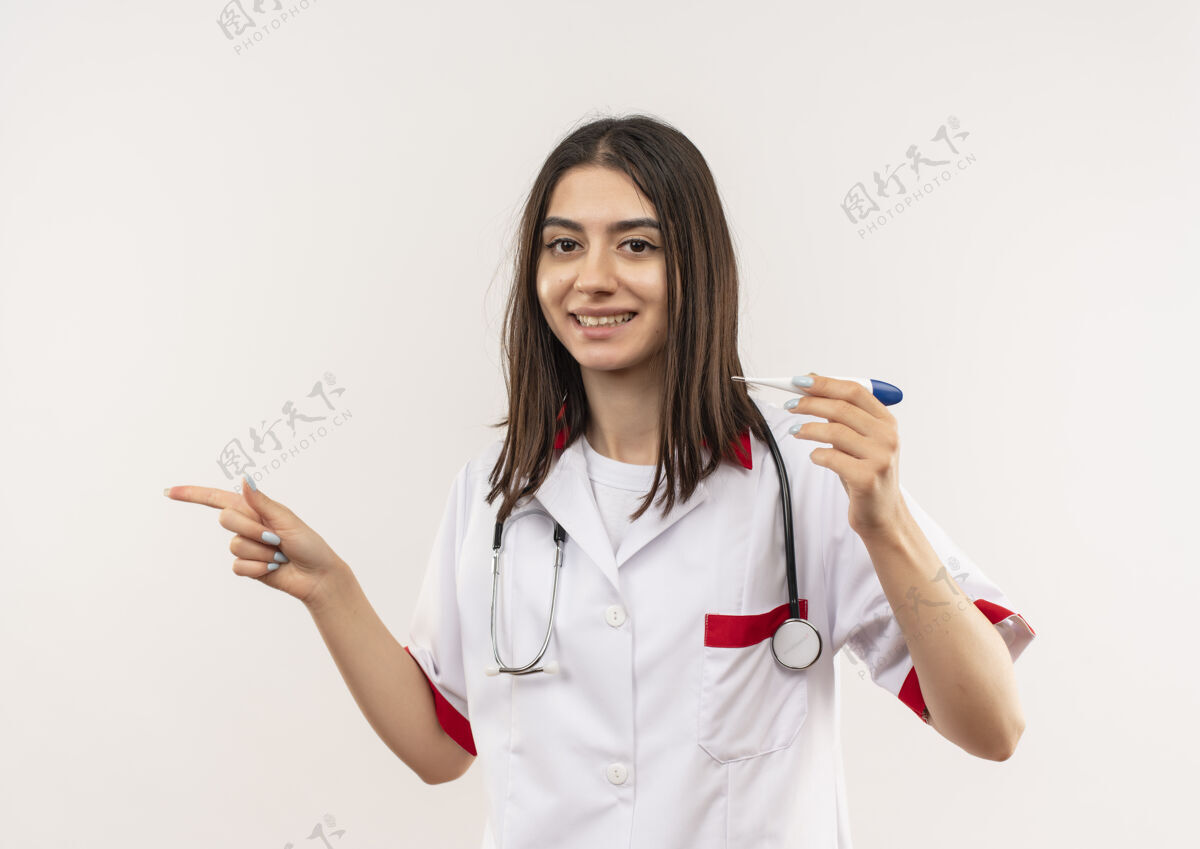 手指身穿白大褂的年轻女医生 脖子上戴着听诊器 拿着数字体温计 手指指向一边 微笑着站在白墙上姿势体温计女人