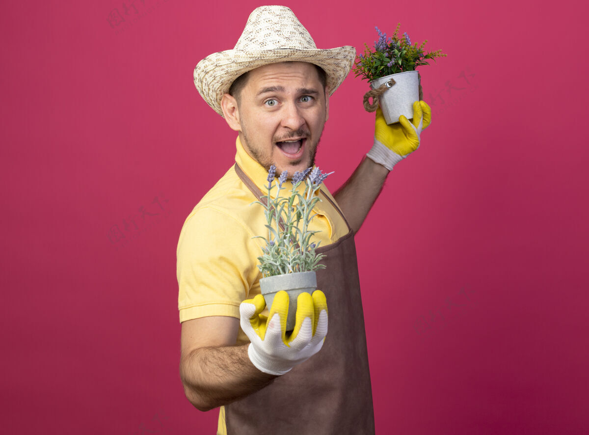 制服年轻的园丁穿着连体衣 戴着帽子 戴着工作手套 手里拿着盆栽植物 站在粉色的墙上 兴高采烈地微笑着看着前方工人看工作