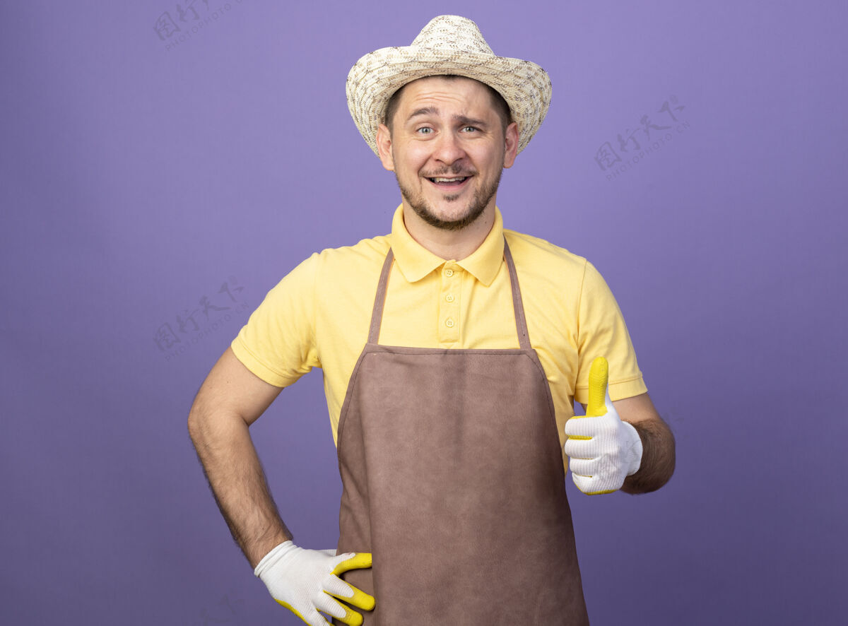 欢呼年轻的园丁穿着连体衣 戴着帽子 戴着工作手套 站在紫色的墙上高兴地微笑着竖起大拇指年轻人穿工作