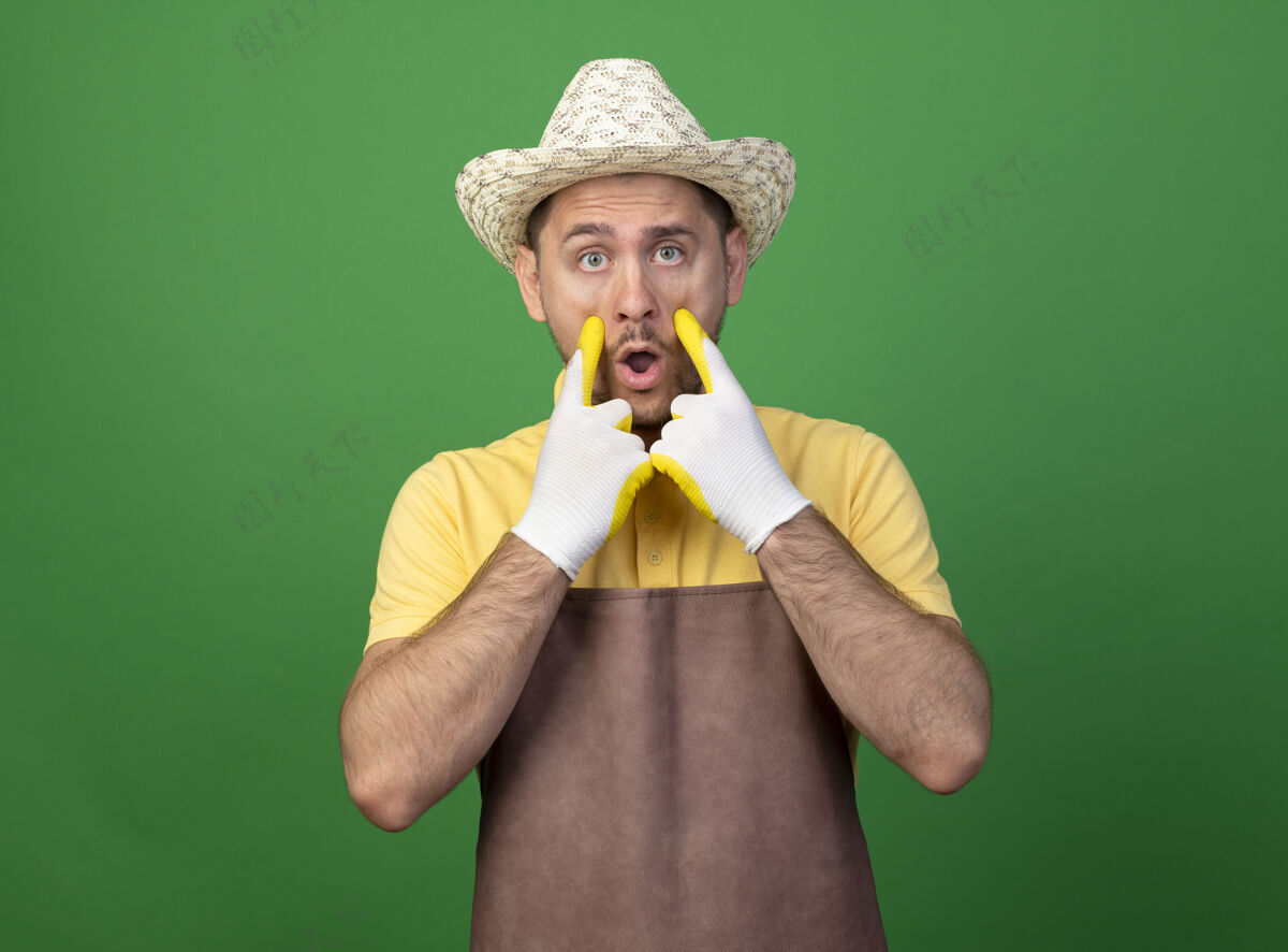 手套年轻的园丁穿着连体衣 戴着帽子 戴着工作手套 站在绿色的墙上 看着他惊讶的眼睛人穿制服