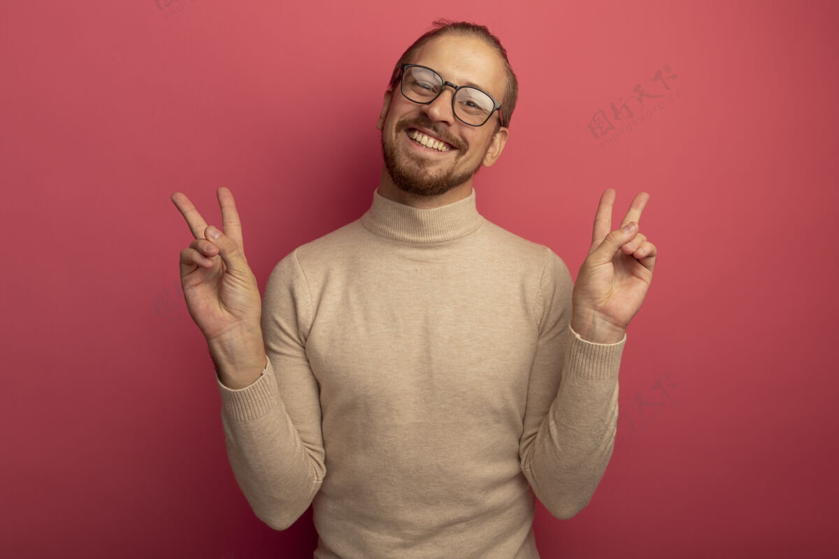 米色快乐的年轻帅哥 穿着米色高领毛衣 戴着眼镜 面带微笑 双手站在粉红色的墙上 露出v字标志人两者表情