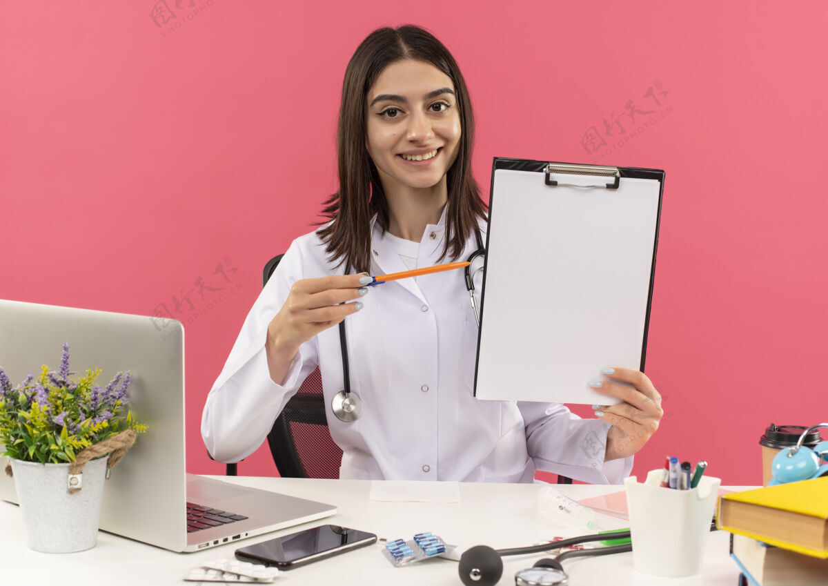 坐着年轻的女医生穿着白大褂 脖子上戴着听诊器 手里拿着空白页的剪贴板 用笔指着剪贴板 微笑着坐在桌边 笔记本电脑盖在粉色的墙上笔脖子医院