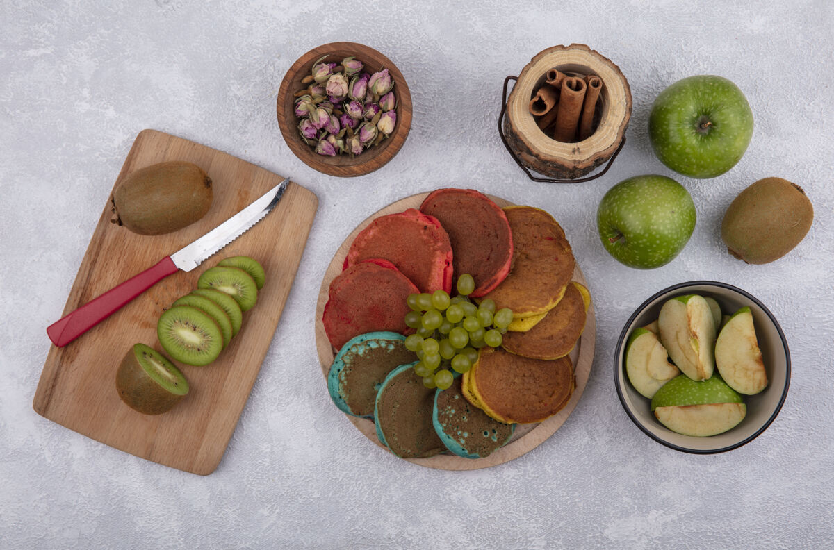 薄煎饼顶视图彩色薄烤饼与绿色葡萄的立场与绿色苹果和奇异果与肉桂在白色背景板刀木板苹果肉桂