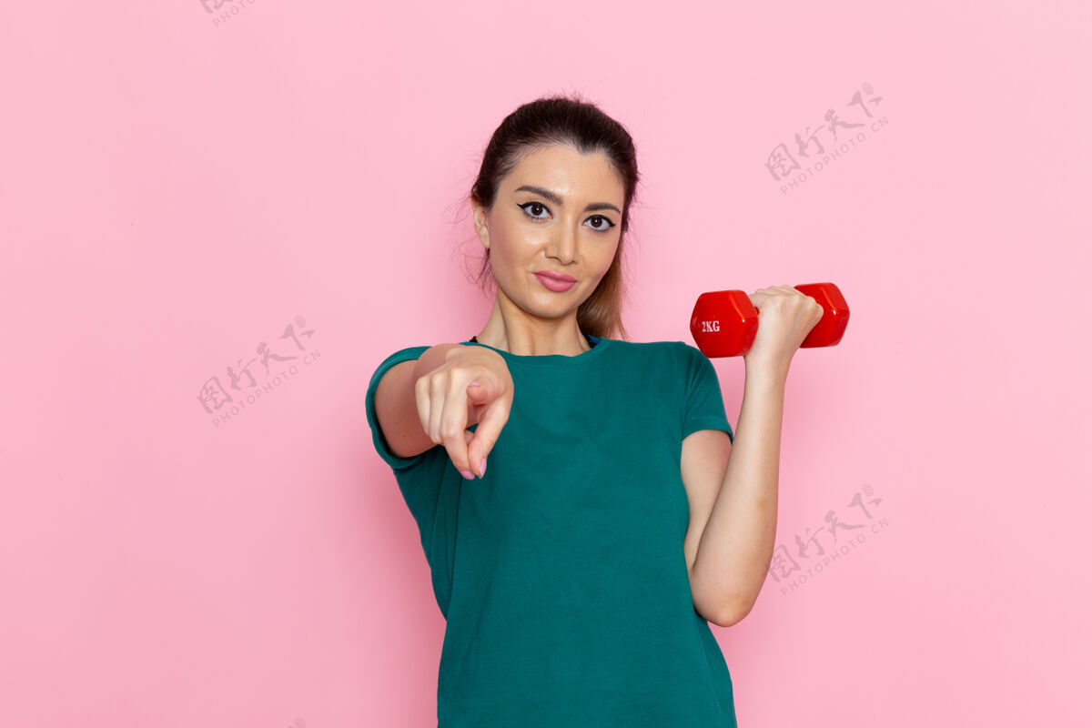 设备正面图年轻女性手持哑铃在粉红色的墙上运动员体育锻炼健康锻炼运动女性体重