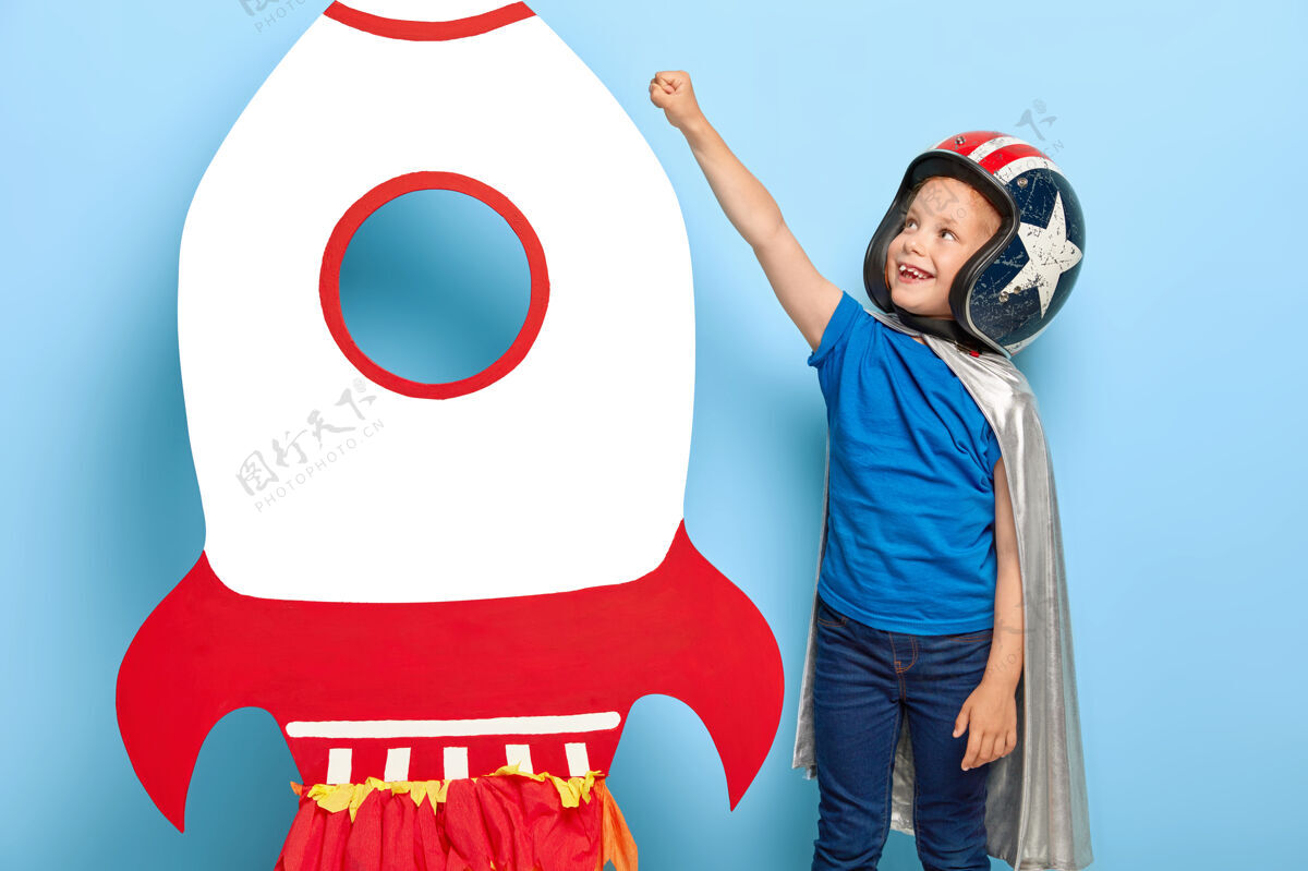 飞行员漂亮的小孩握紧拳头 做飞翔的手势 在玩具火箭附近摆姿势头饰极限幼儿园