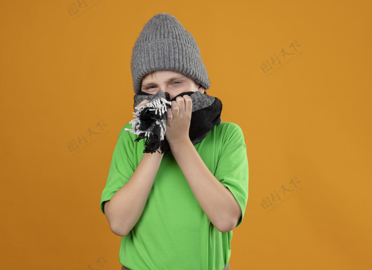 温暖生病的小男孩穿着绿色t恤 戴着暖和的围巾 戴着帽子 戴着围巾 站在橙色的墙上感觉不舒服盖站立少