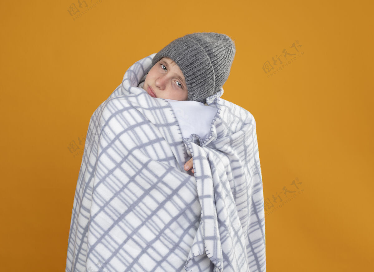 穿生病的小男孩戴着暖和的帽子 裹着毯子 站在橙色的墙上发烧 感觉不舒服包裹疾病男孩