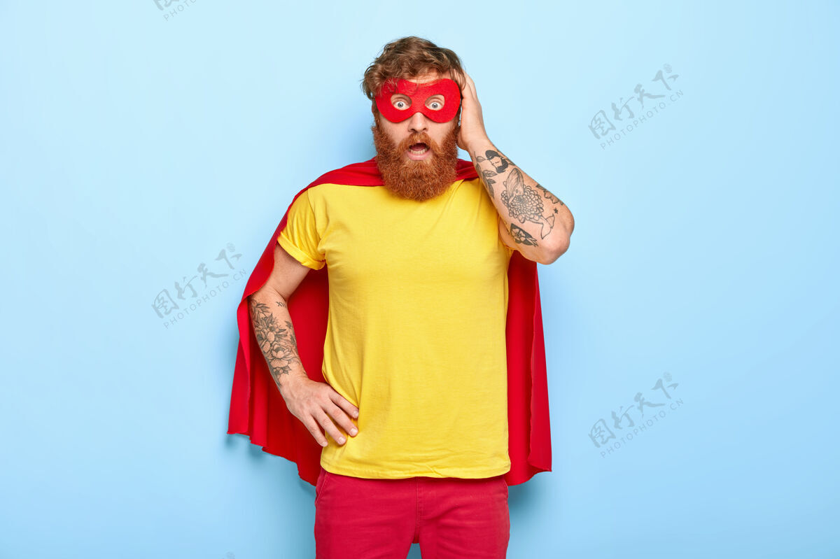纹身惊恐的红发男子惊恐地看着镜头 假装在与邪恶战斗 穿着超级英雄的衣服力量斗篷凝视