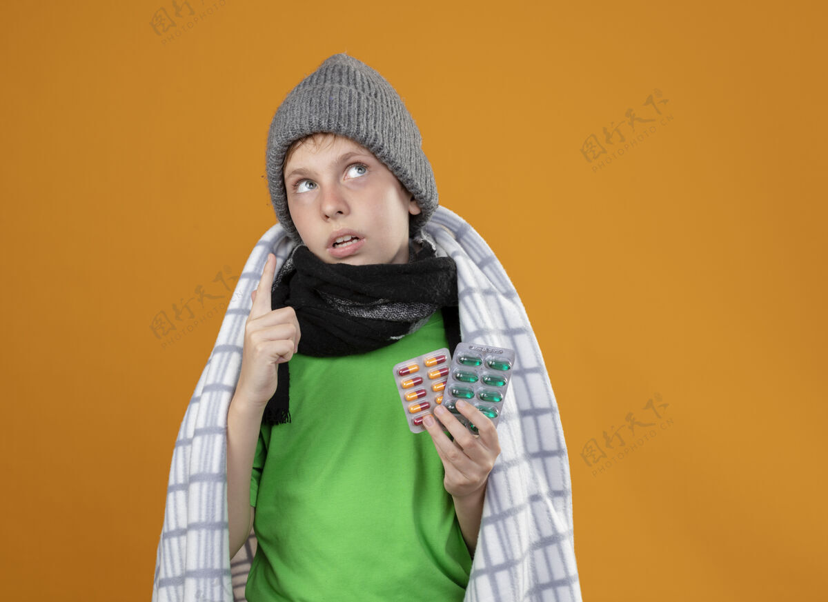 包裹生病的小男孩戴着暖和的帽子 围着围巾 裹着毯子 发烧 服用避孕药 身体不适 抬起头来 用手指指着橙色的墙壁帽子发烧男孩