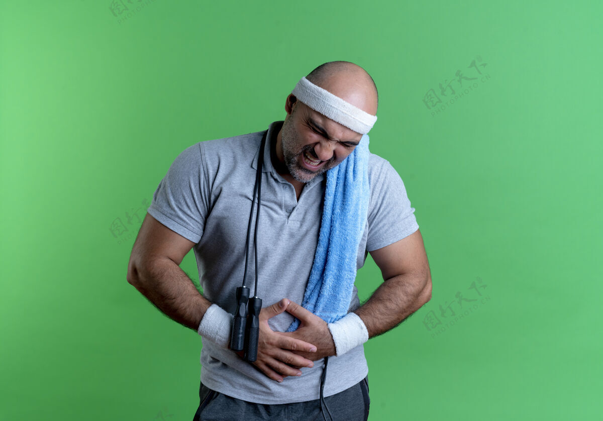 脖子戴着头巾 脖子上缠着毛巾 站在绿墙上摸着肚子的成熟运动型男人毛巾站教练