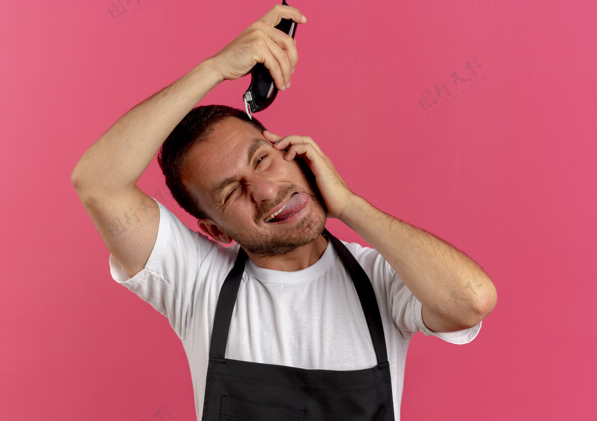 理发师穿着围裙的理发师站在粉红色的墙上 用修剪器伸出舌头刮头发男人工人围裙