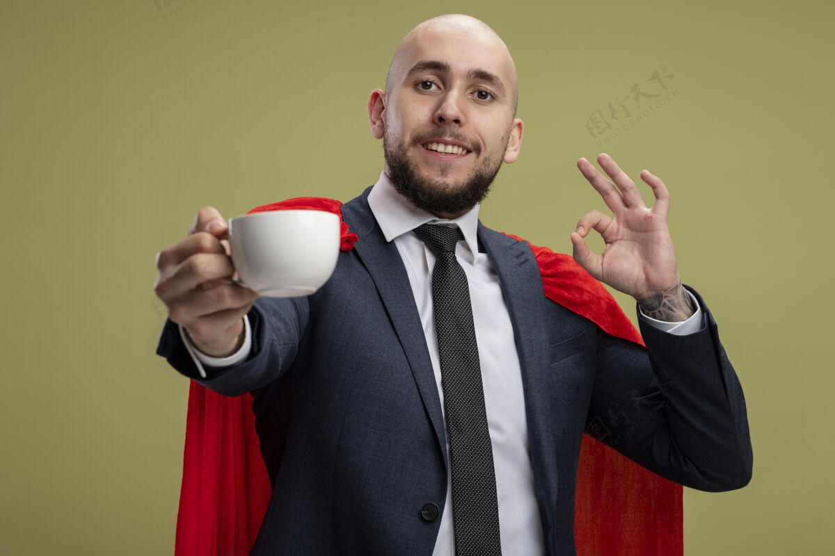 微笑超级英雄 留着胡子 披着红斗篷的商人 手里拿着一杯咖啡 微笑着 在浅绿色的墙壁上立着ok标志秀超级英雄杯子