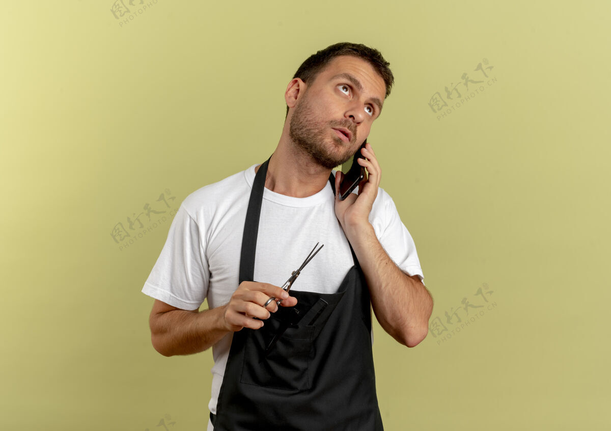 男围裙上的理发师拿着剪刀讲着手机 脸上带着严肃的表情站在轻墙上围裙工作手机