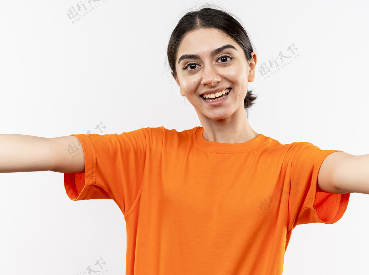 朋友穿着橙色t恤的年轻女孩友好地微笑着 双手站在白色的墙上做欢迎的手势女孩站微笑