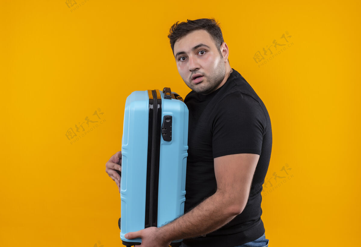 抱着身穿黑色t恤的年轻旅行家拿着手提箱 站在橙色的墙上吓坏了旅行人年轻人