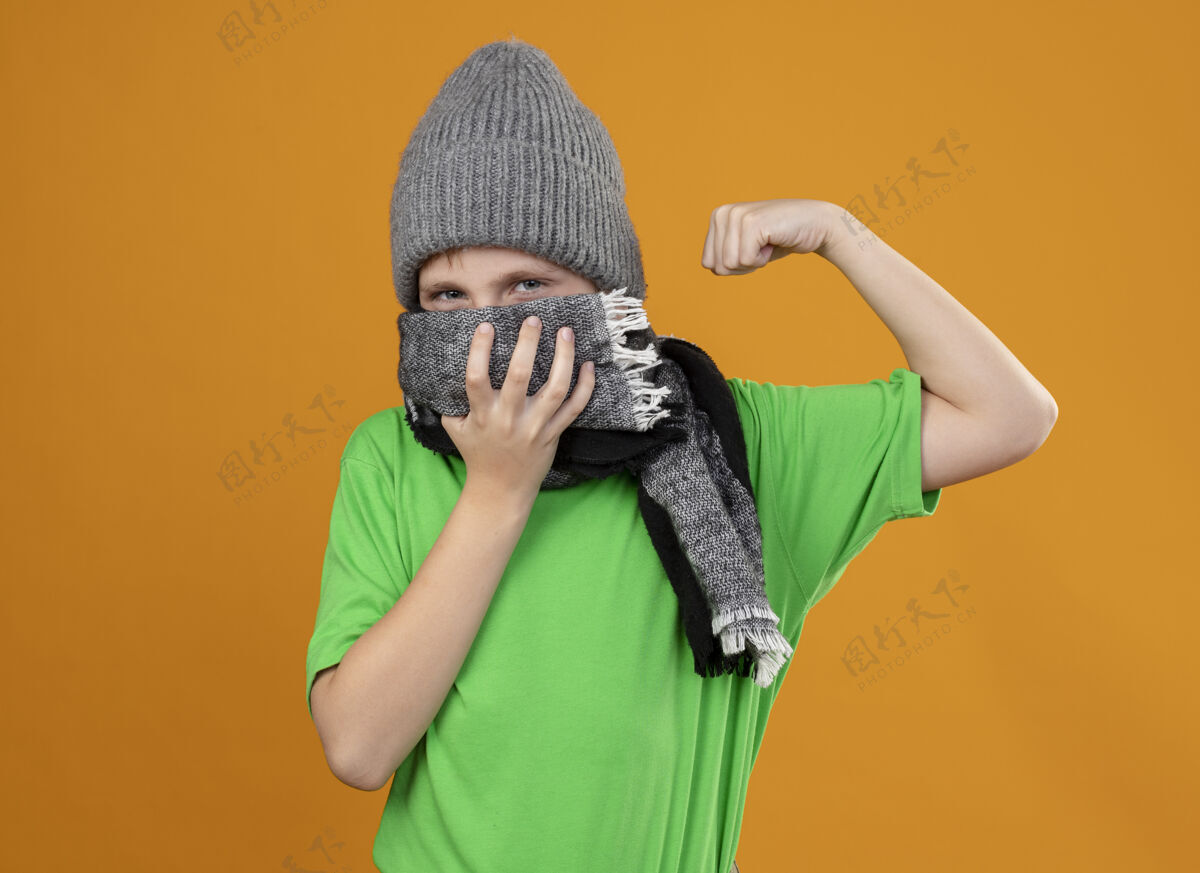 提高生病的小男孩穿着绿色t恤 戴着暖和的围巾 举着帽子 站在橙色的墙上感觉好多了穿小男孩