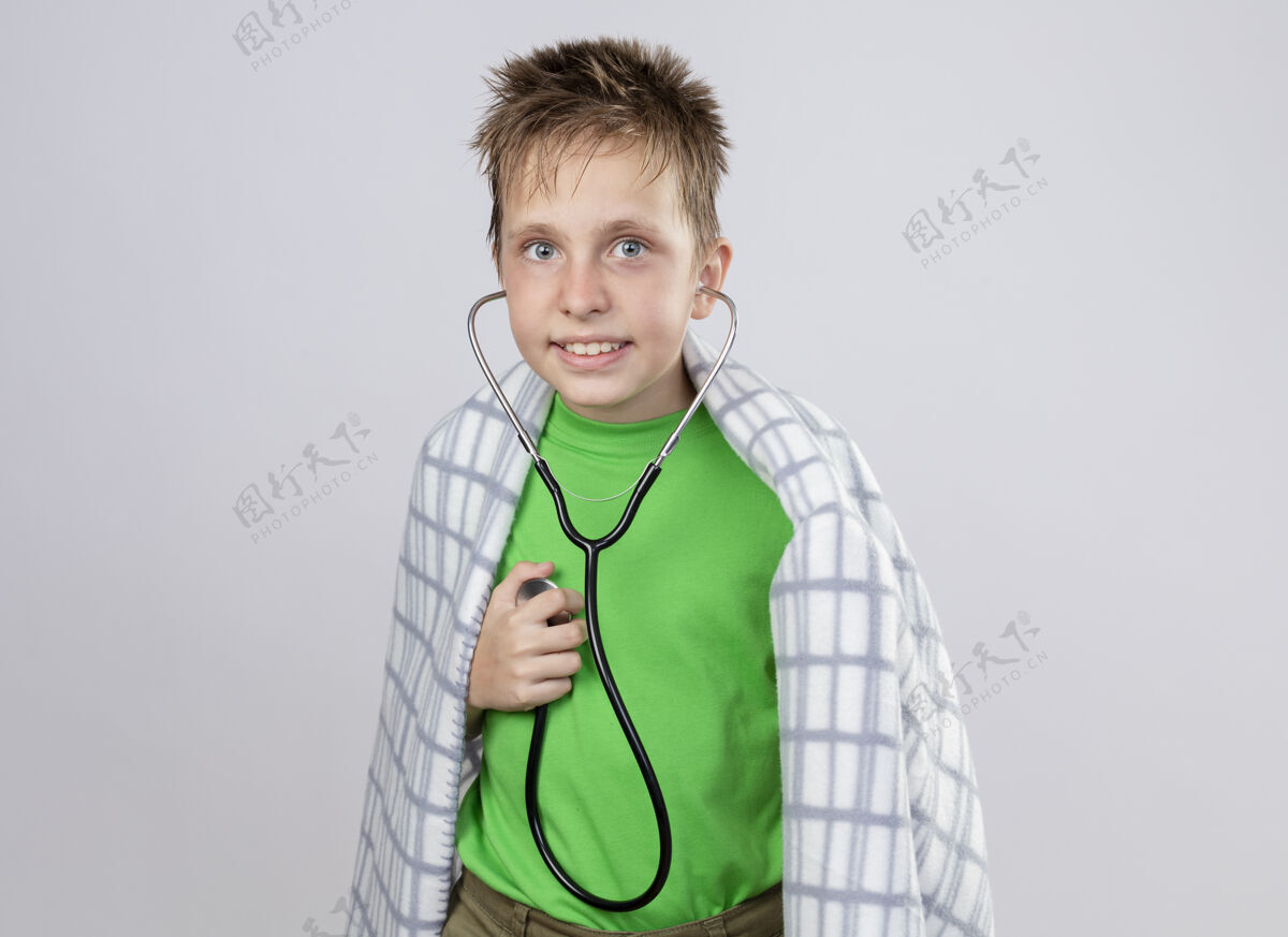 听力生病的小男孩穿着绿色t恤 裹着毛毯 脖子上戴着听诊器 站在白色的墙上 听着心跳混乱听诊器男孩围绕