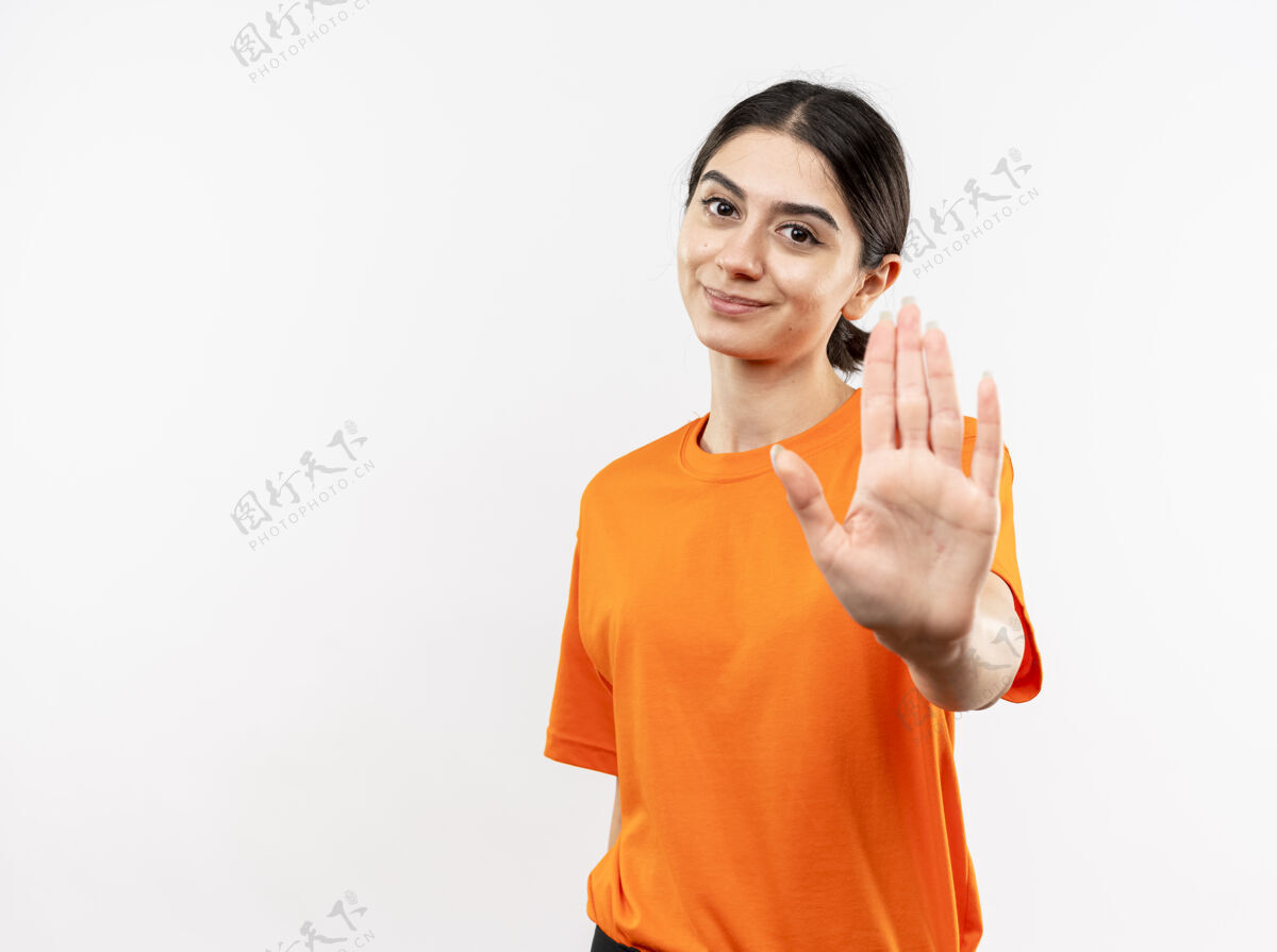 年轻穿着橙色t恤的年轻女孩站在白色的墙上 微笑着用手做着停下来的手势看做站