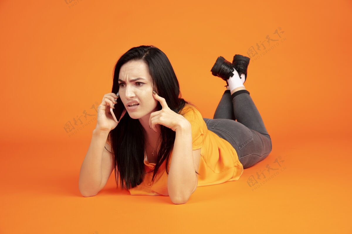 女商人橙色工作室背景上的白种人年轻女子肖像穿着衬衫的漂亮黑发女性模特人类情感的概念 面部表情 销售 广告广告空间电话交谈年轻面部员工