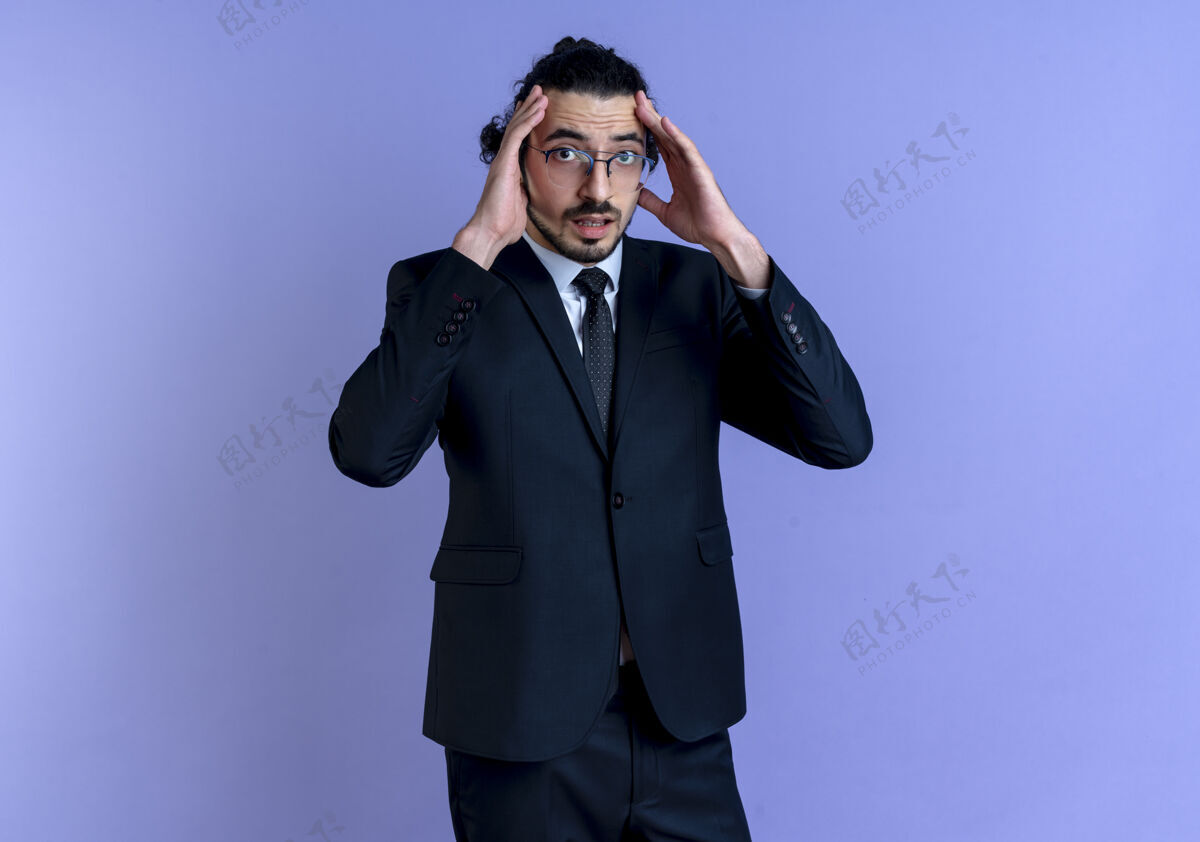 领带一个穿着黑色西装戴着眼镜的商人 两手站在蓝色的墙壁上 两手靠近头部 两手困惑地看着前方西装优雅公民