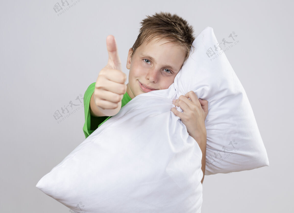 拇指生病的小男孩穿着绿色t恤感觉好多了抱着枕头看着竖起大拇指微笑着站在白色的墙上微笑站显示