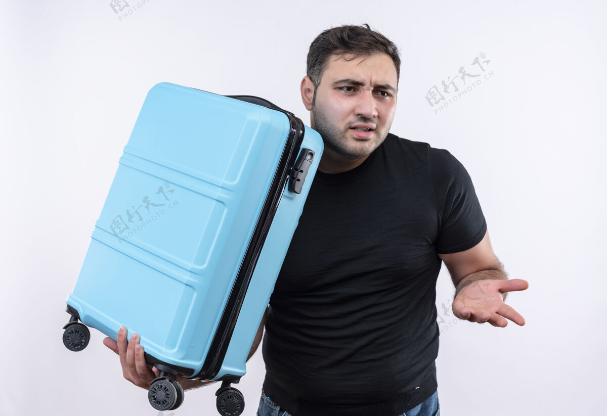 焦虑穿着黑色t恤的年轻旅行家拿着手提箱 站在白色的墙上 困惑地 焦急地看着一边困惑旅行者手提箱