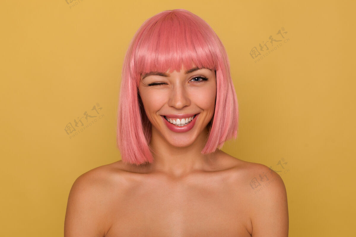 特写特写镜头：迷人欢快的年轻粉红头发女士 留着时髦的短发 在芥末墙上摆着姿势 顽皮地看着镜头 眨眨眼休闲短裤20多岁
