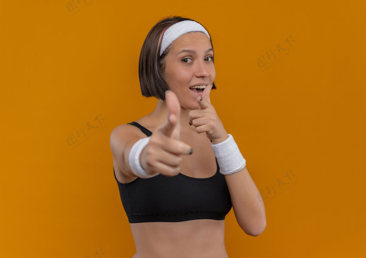 人身着运动服的年轻健身女士 头戴着积极的头巾 用食指指着站在橘色墙上的摄像机 高兴极了健身运动员积极