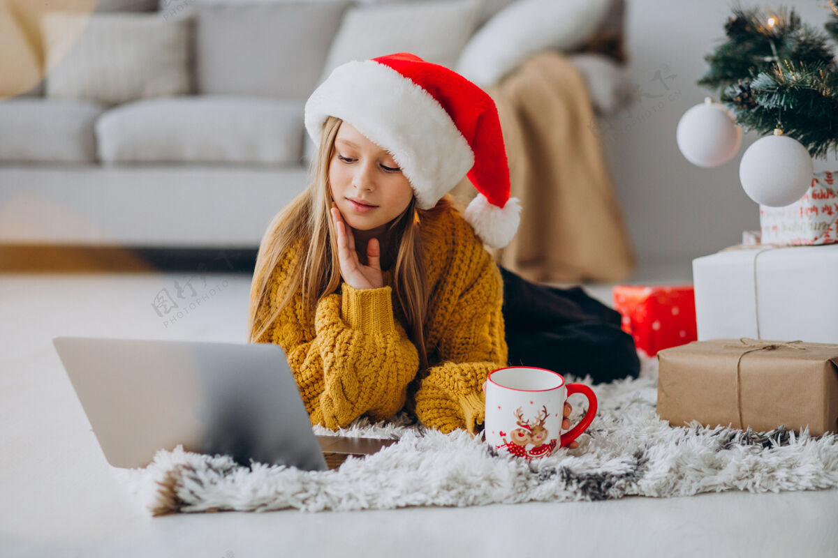 圣诞销售可爱的女孩在圣诞树旁用电脑冠状病毒圣诞礼物现代
