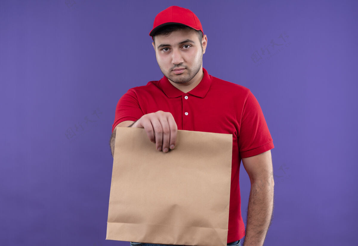 姿势年轻的送货员穿着红色制服 戴着帽子 手里拿着纸包 严肃的脸站在紫色的墙上公民拿着人