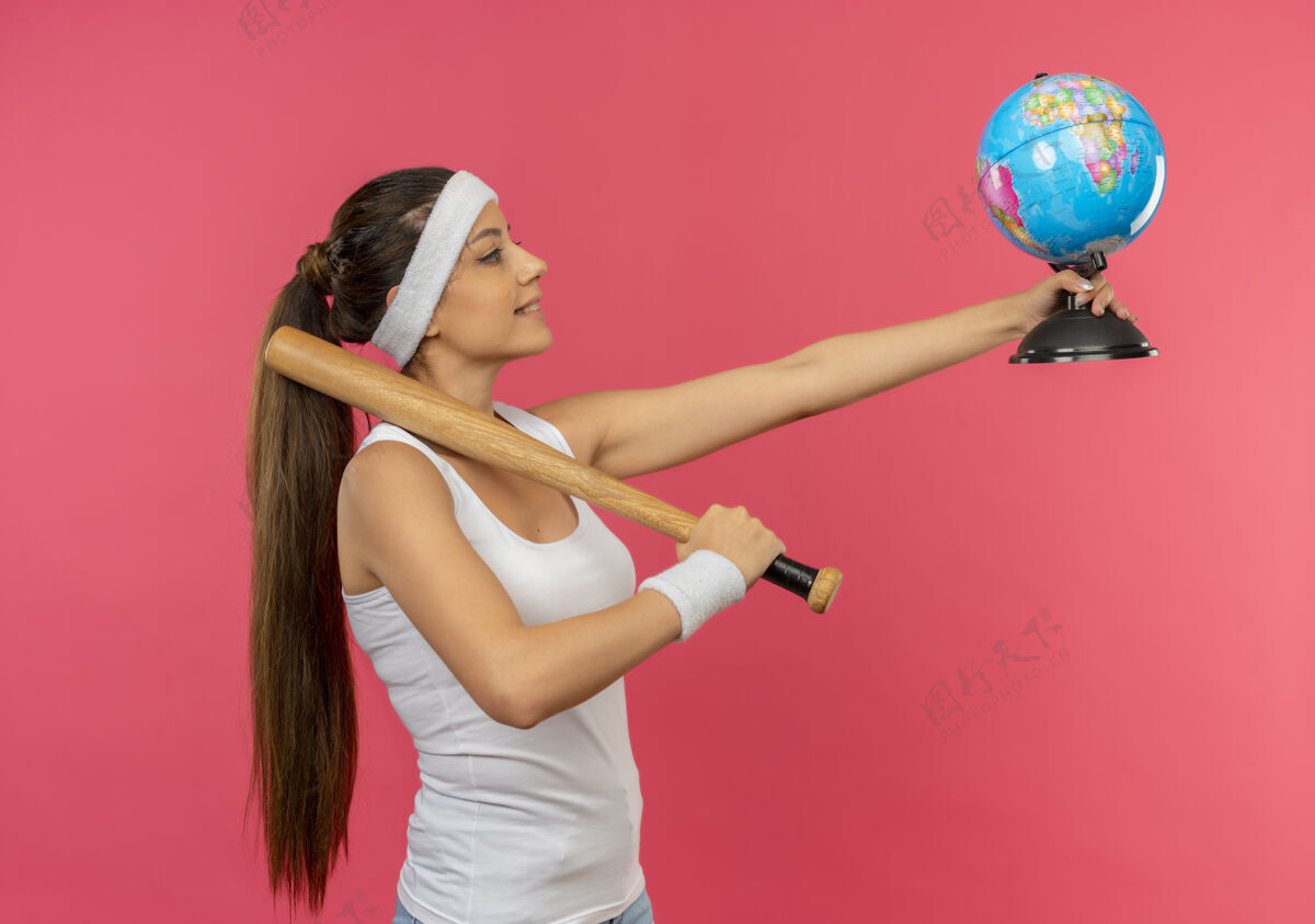 人身穿运动服 头箍手持球棒 面带微笑的年轻健身女士自信地站在粉色的墙上公民头带抱着