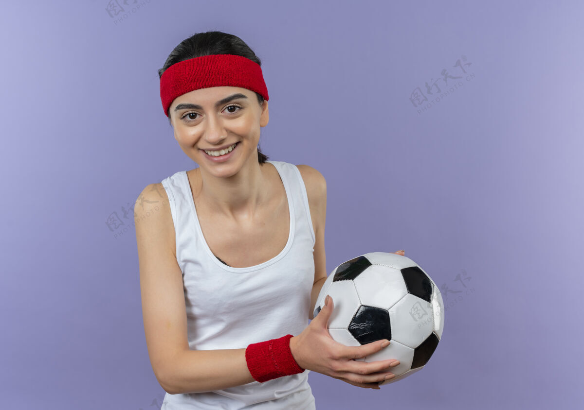 人身穿运动服 头戴钢带 面带微笑地拿着足球站在紫色墙壁上的年轻健身女士健康运动脸