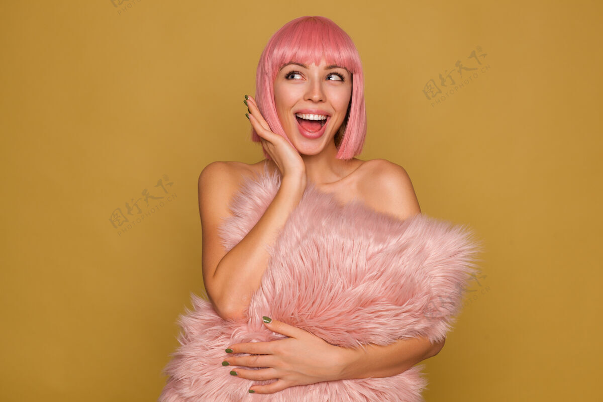 自然快乐的年轻迷人的粉红色头发的女性 绿色的指甲 愉快地看着旁边 张大嘴巴 拿着蓬松的枕头放在芥末墙上年轻女性肩膀
