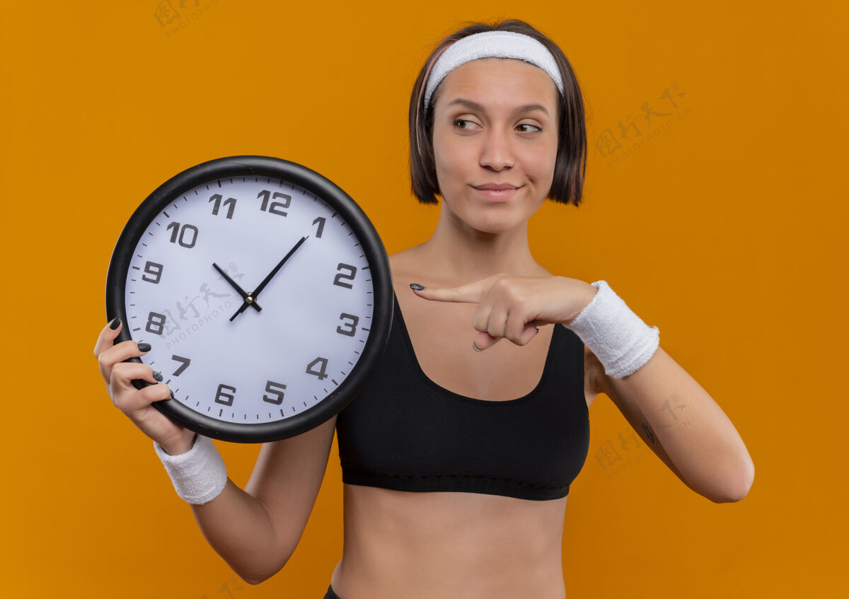 人身穿运动服的年轻健身女士 戴着头带 拿着挂钟 手指指着挂钟 脸上带着自信的微笑站在橙色的墙上手指年轻人微笑