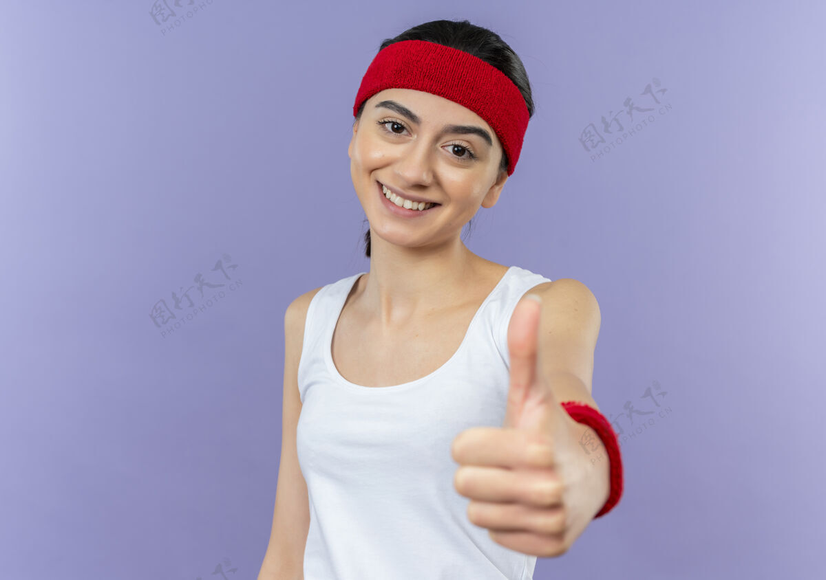 姿势身穿运动服 头箍微笑的年轻健身女士站在紫色的墙上 愉快地竖起大拇指人拇指运动装