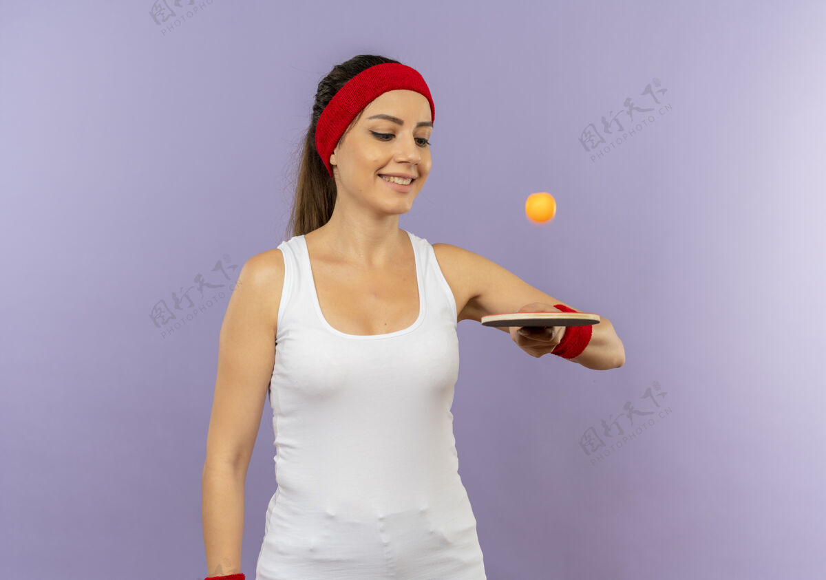 市民身穿运动服的年轻健身女 头戴花环 手持球拍和乒乓球 站在灰色的墙壁上欢快地微笑着运动装健身网球