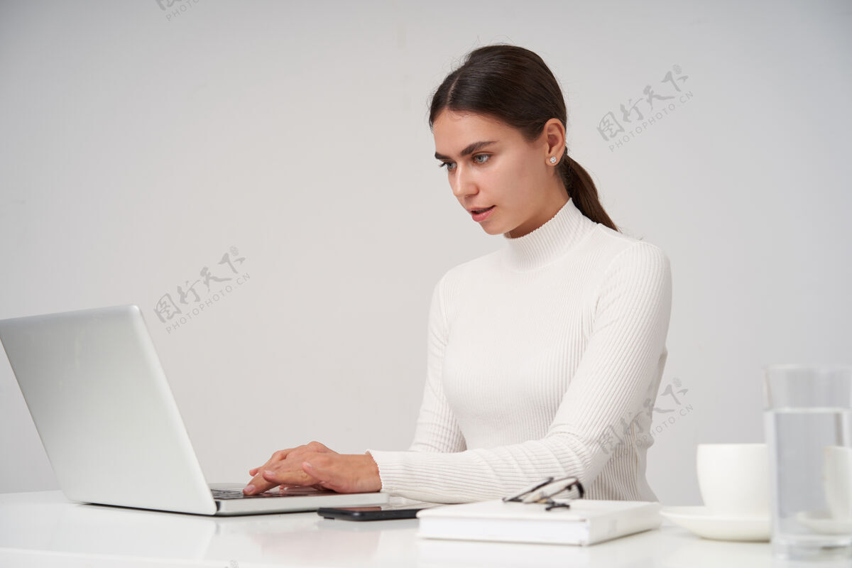 欧式身着白色针织圆领衫的黑发女商人坐在白色墙壁上 手里拿着笔记本电脑 手放在键盘上 认真地看着屏幕女士马尾辫手