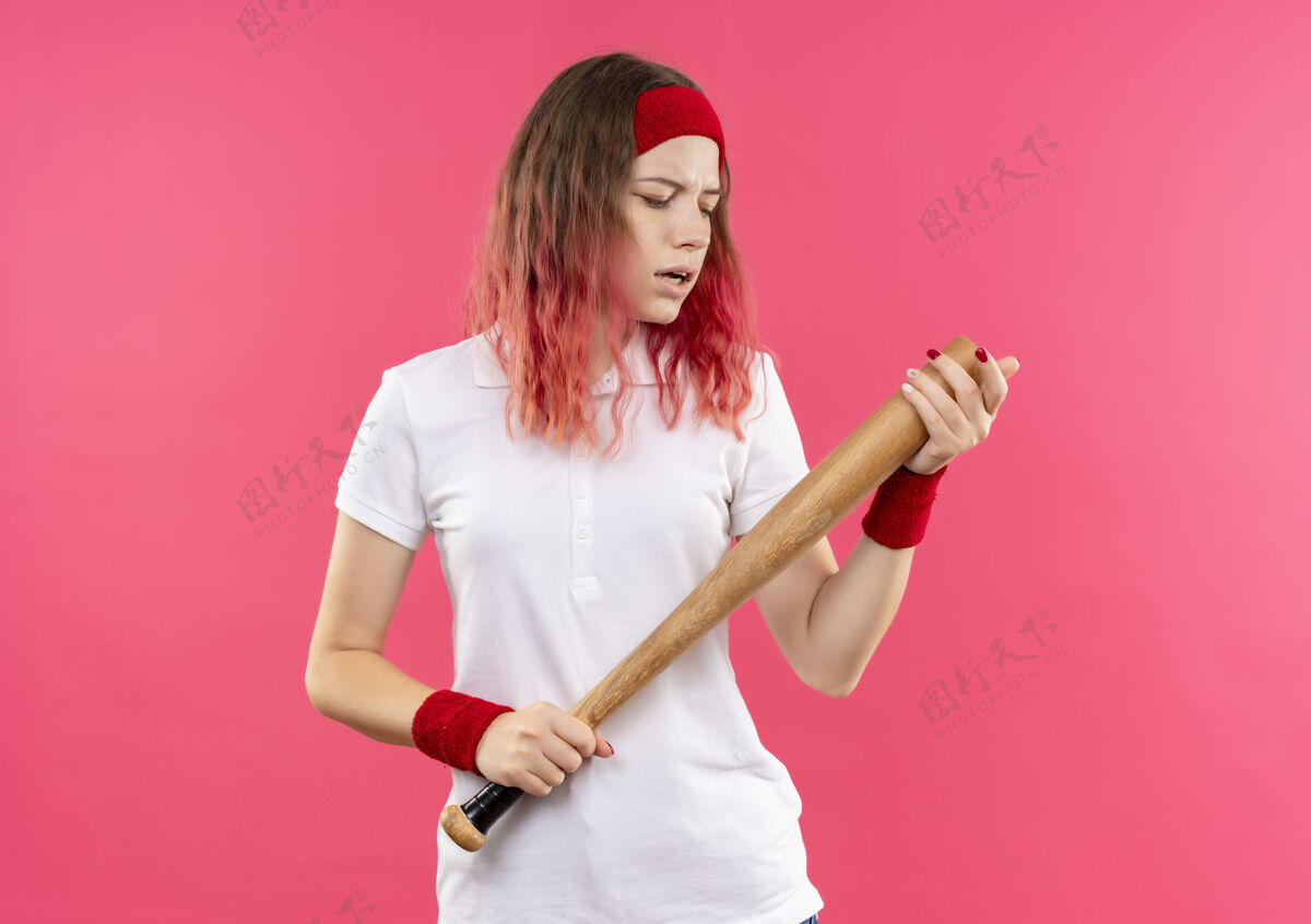 运动戴着头巾的年轻运动型女子拿着球棒去打棒球 站在粉红色的墙上显得很自信女人蝙蝠人