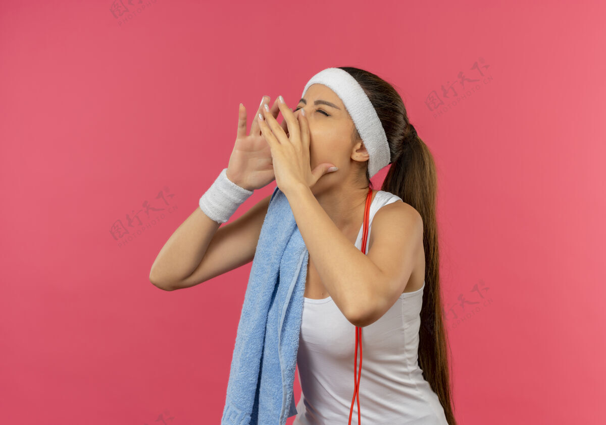 人身穿运动服的年轻健身女 头上戴着头带 肩上扛着毛巾 手靠嘴站在粉红色的墙上大声喊叫嘴市民近