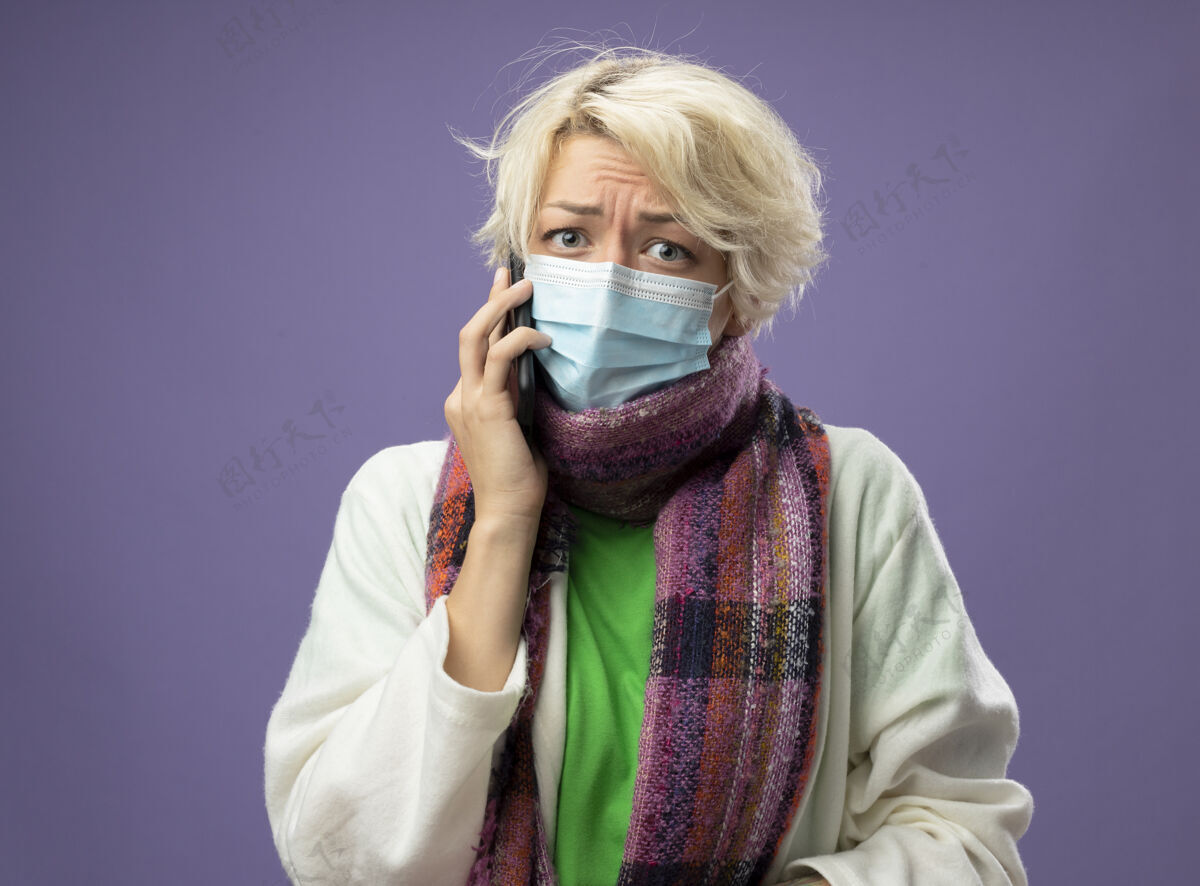 短裤生病的不健康的女人 短头发 戴着暖和的围巾和面罩 站在紫色的背景上讲手机 看起来很紧张 很担心生病保护温暖