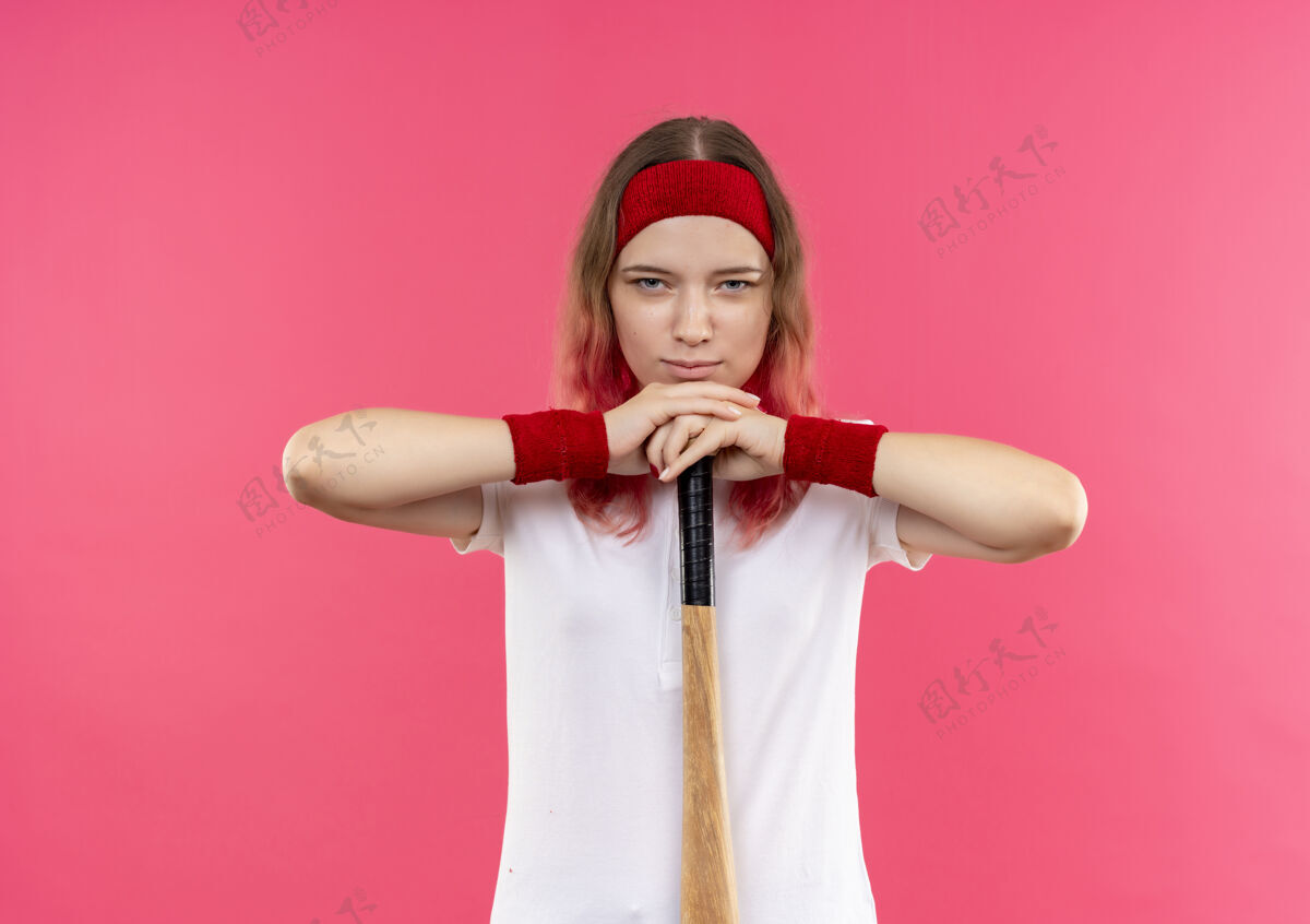 头带戴着头巾的年轻运动女郎拿着球棒去打棒球 自信的表情站在粉色的墙上运动员人运动