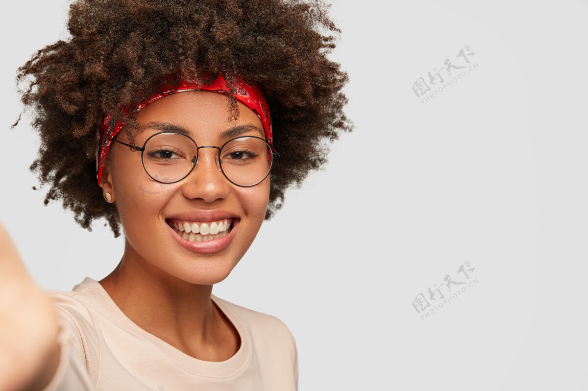 头像特写镜头快乐的女性女孩有着牙齿般的微笑 非洲式的发型 漂亮干净的皮肤 戴着透明的眼镜 伸手拿着不可识别的装置 在白色的墙壁上自拍快乐长相外表