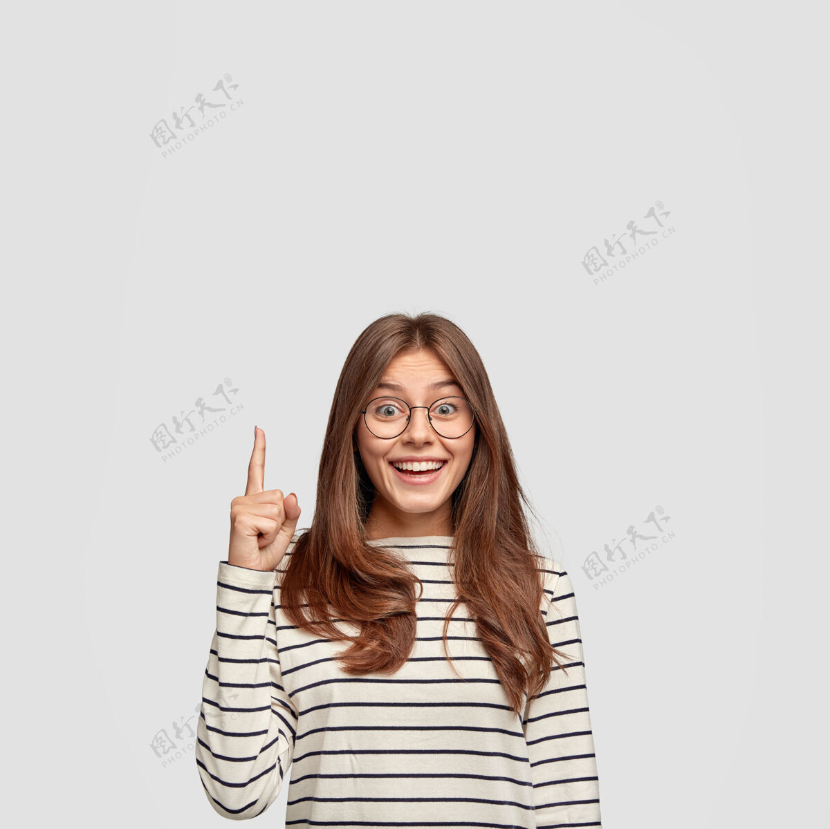 手指独立拍摄的开朗白种女人直发 展示自由空间 用食指点出你的广告内容 穿着休闲条纹毛衣 隔着白墙眼镜眼镜白种人