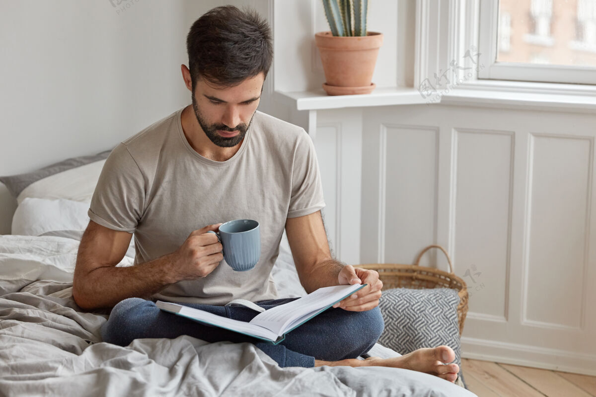 卧室认真专注的男性在休息日看书 参与阅读 喝热饮料 盘腿坐在床上 穿休闲t恤和裤子人 知识 教育 休闲胡须放松识字