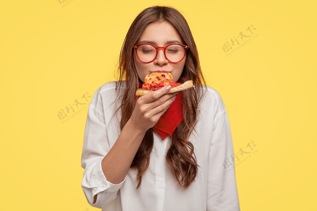 味道嗯 那么美味的黑发美女吃意大利披萨片 闭上眼睛不高兴 享受美味 戴着眼镜和衬衫 隔离在黄色的墙壁上吃的概念披萨店热的快餐