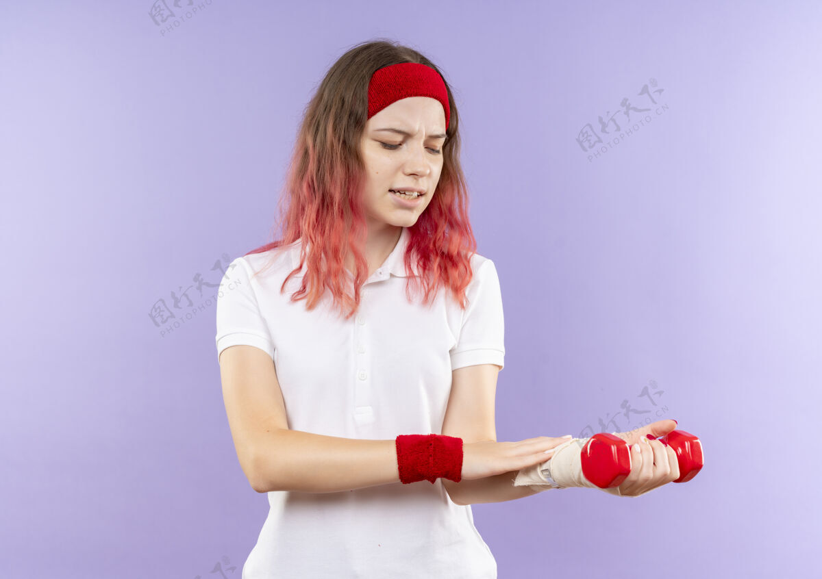 抱着年轻的运动女子举着两个哑铃做运动看着她缠着绷带的手腕站在紫色的墙上感到疼痛疼痛运动运动服