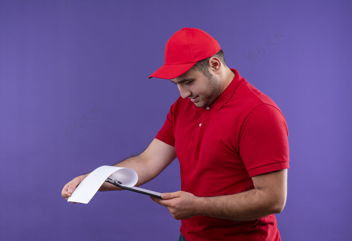 男人年轻的送货员穿着红色制服 戴着帽子 看着剪贴板上的空白页 微笑着自信地站在紫色的墙上帽子公民年轻